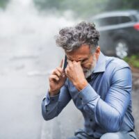 Mature man making a phone call after a car accident, smoke in the background.